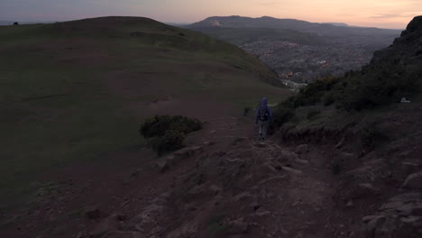 Dolly-follow-shot-of-girl-walking-down-the-Arthurs-seat-mountain-on-the-rocky-hiking-trail-in-evening,-dusk-with-city-of-Edinburgh-in-the-background-during-wonderul-blue-hour