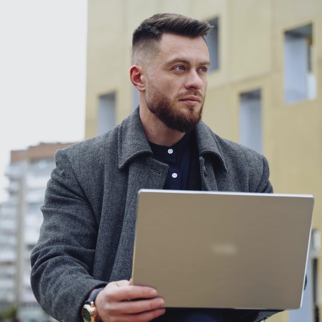 Portrait of a handsome man with a laptop. Young bearded businessman in a coat works on a wireless gadget on the urban background outdoors.