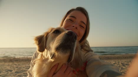 View-from-the-side-of-the-phone,-a-blonde-girl-and-her-light-dog-take-a-selfie-on-the-background-of-a-sunny-beach-in-the-morning,-close-up-shot