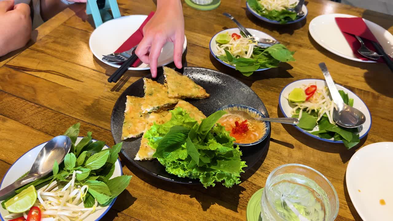 Hands picking crispy pancake from a Vietnamese dish on a wooden table, surrounded by fresh greens and dipping sauce