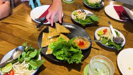 hands picking crispy pancake from a vietnamese dish on a wooden table, surrounded by fresh greens and dipping sauce