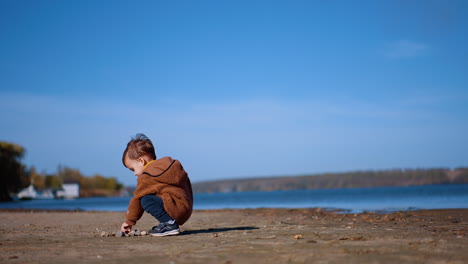 caucasian toddler comes up to a pile of stones on the ground. kid takes a stone and runs to water to throw it. low angle view.