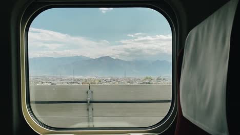 Vista-De-Una-Ciudad-Con-Montañas-En-El-Fondo-En-Un-Día-Soleado-Desde-Una-Ventana-De-Tren-Mientras-Va-Rápido