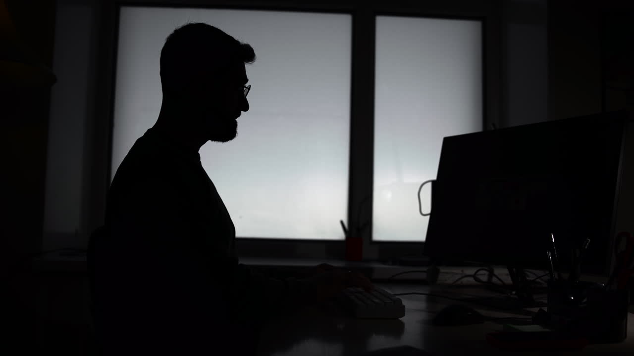Silhouette di uomo chinato sul laptop vicino a una finestra retroilluminata, postura contemplativa e profilo ad alto contrasto. Media