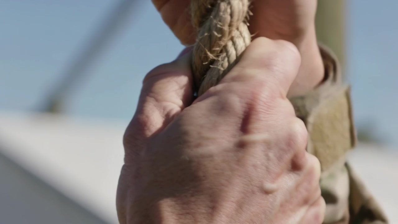 Mani che afferrano una manica mimetica tirando una corda, la mano destra che afferra la sinistra unendosi per stabilizzare su una struttura esterna.