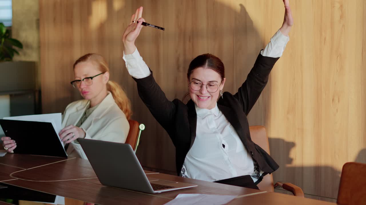 счастливая брюнетка в круглых очках крутится на стуле за столом в офисе в то время как борется за успех и победу за своим столом в современном офисе