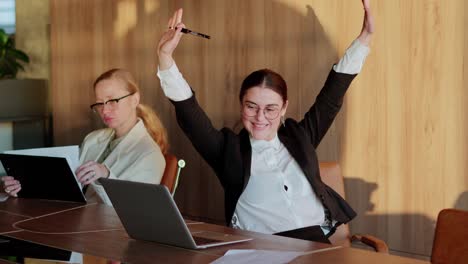 Happy-brunette-businesswoman-in-round-glasses-spins-on-a-chair-at-a-desk-in-the-office-while-fighting-for-success-and-victory-at-her-desk-in-a-modern-office