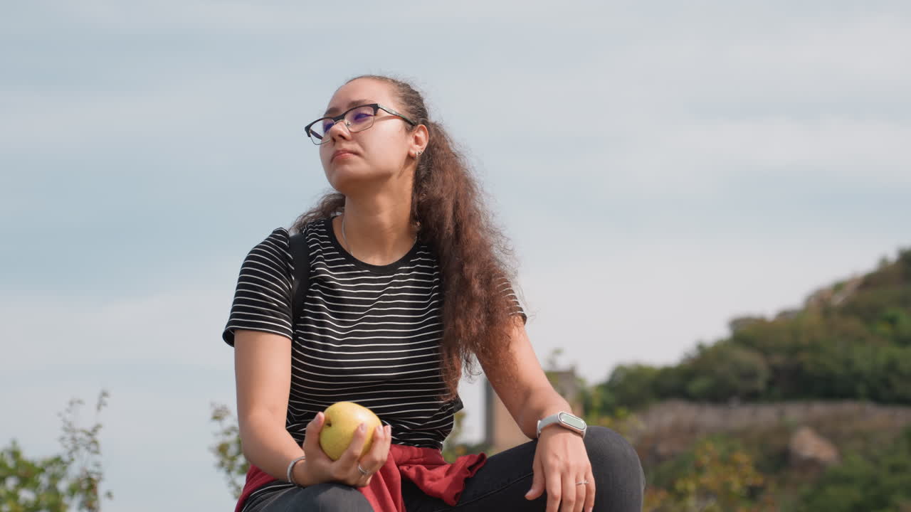 Giovane donna che porta una mela alle labbra, profilo pensieroso contro il cielo nuvoloso, camicia a righe e occhiali, zaino a fianco, rovine di una fortezza sullo sfondo, stato d'animo riflessivo che assapora lo spuntino durante l'escursione.