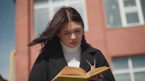 close-up of student flipping pages of a book while wind flutters her hair, set against a red brick building background, showcasing a thoughtful reading moment outdoors under bright sunlight