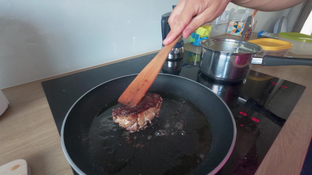 A hand carefully sears a thick, seasoned steak in a frying pan over an induction stove in a modern kitchen. Close-up shot highlights rich textures, sizzling oil, and warm daylight ambiance.
