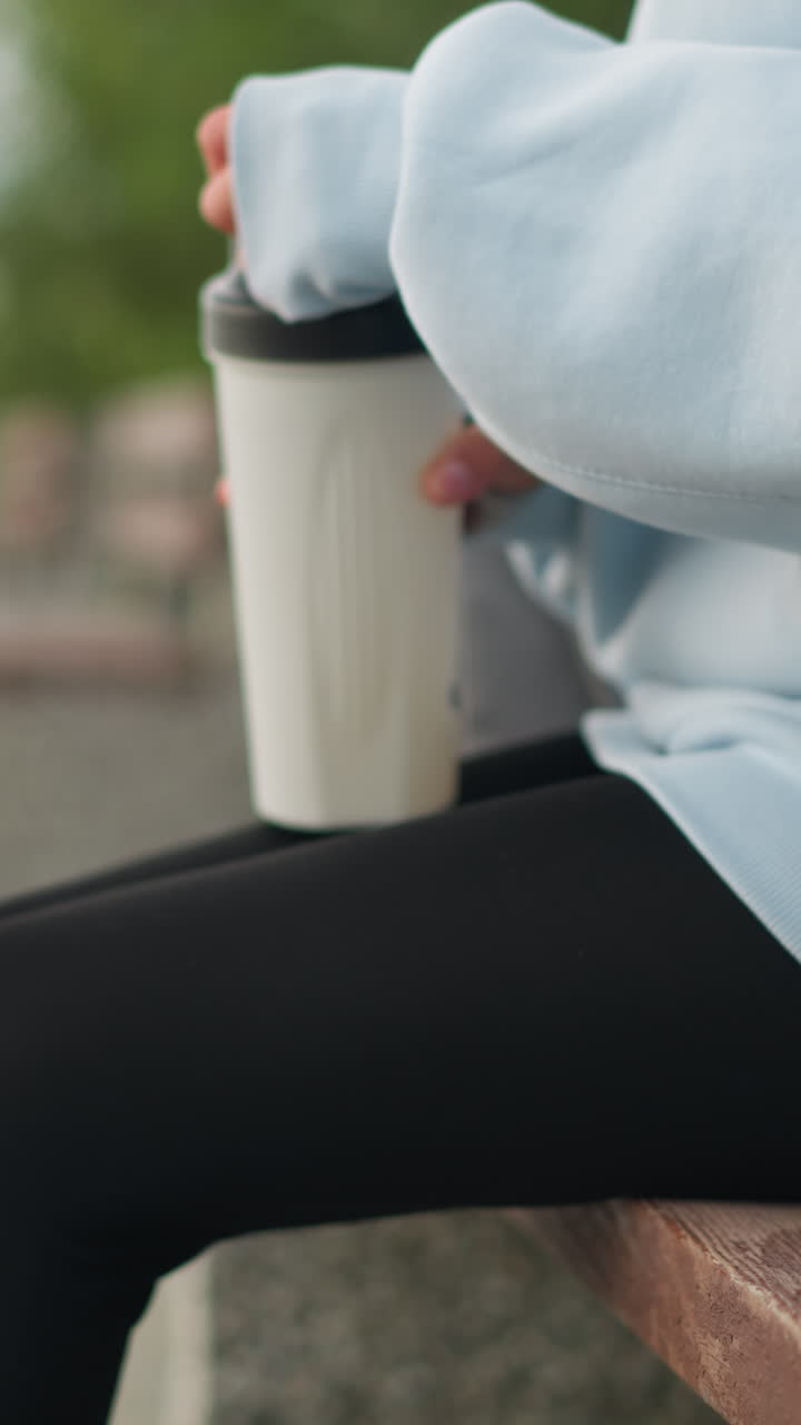 Luftfoto af en kvinde, der holder en vandflaske og åbner den, mens hun sidder på en bænk i en park, med sløret baggrund, fokuseret på hydrering og afslapning, perfekt til velvære- og naturøjeblikke