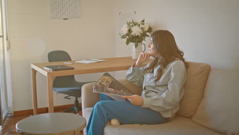 thoughtful model resting sofa holding magazine at sunlight home. woman relaxing