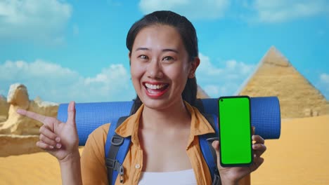 close up of asian female hiker with mountaineering backpack showing mock up green screen smartphone and pointing to side while traveling in pyramid of giza