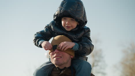 a close-up of a father carrying his young son on his shoulders, as the playful child covers his father's face with a beanie, with blurred view of trees behind them