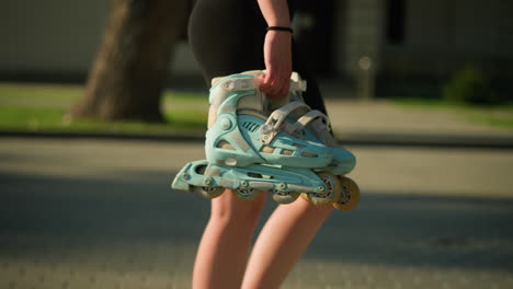 side view of lady walking outdoors holding cyan roller skates in her right hand, with a blurred background featuring greenery, trees, and a building