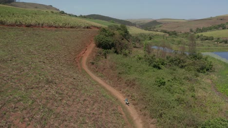 aerial view closely tracking enduro dirt bike rider on sandy trail through crop fields