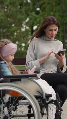 little girl with disability watches cartoons while woman checking social media on smartphone. mother and daughter in wheelchair use devices in park