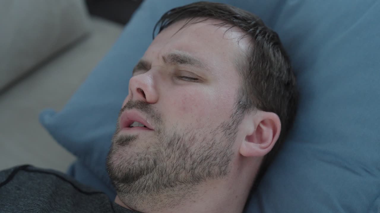 Känslosamt stockfilmmaterial som visar en sjuk man som ligger på en soffa, svettas och ser orolig ut på grund av feber, vilket symboliserar sjukdom, obehag, utmattning och de fysiska effekterna av infektion i hemmet