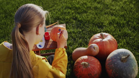 vista trasera: un niño pinta una calabaza, prepara decoraciones para halloween. se sienta en el césped en el patio trasero de la casa
