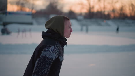 side view boy gliding smoothly on outdoor ice ring wearing patterned winter jacket with hood at sunset golden reflection on icy surface knee bend blurred stadium seating background crisp winter air