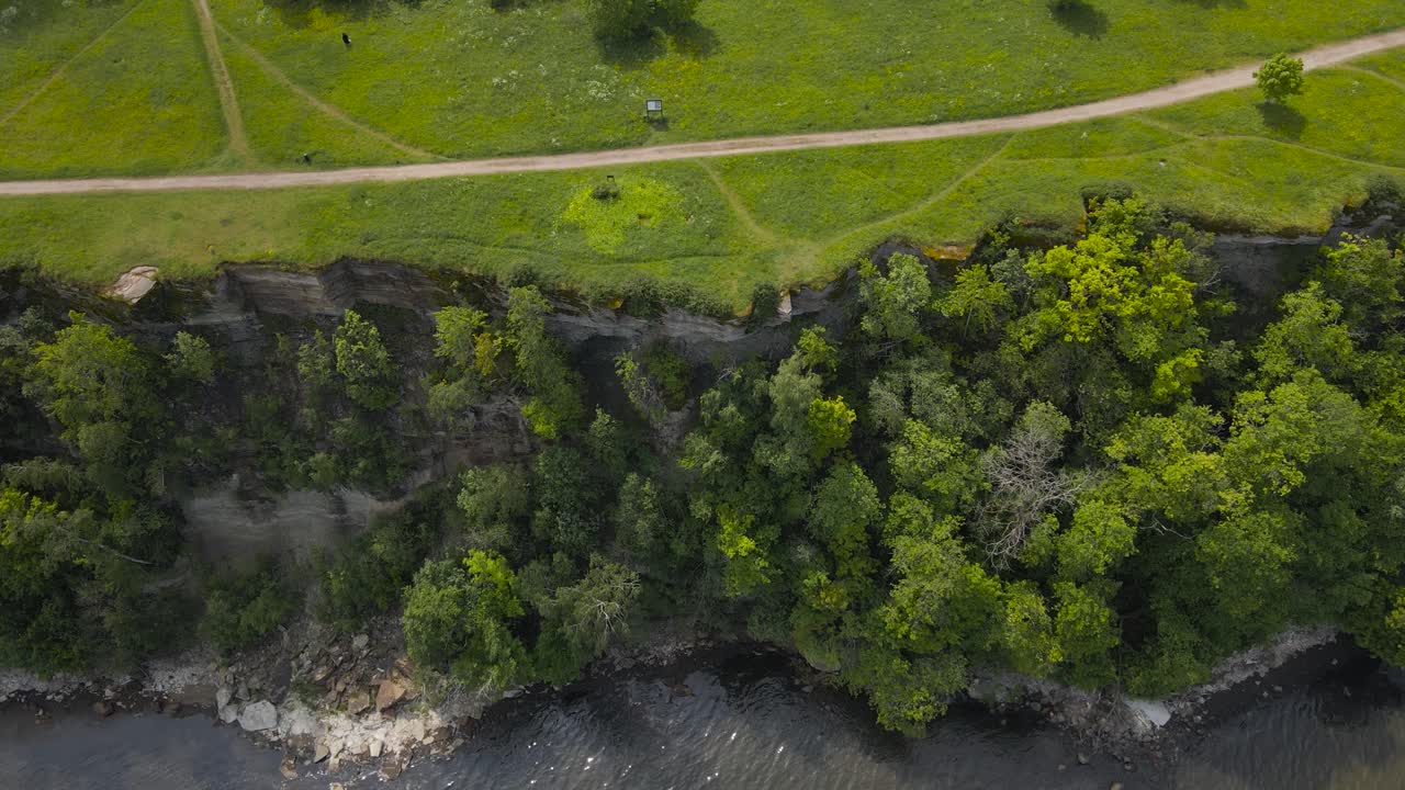 Top down revealing aerial footage showing a steep and high altitude cliff bank in Tabasalu during a sunny summer day. The drone flies backwards reveals the forest and grassy landscape on top of it.