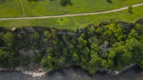 top down revealing aerial footage showing a steep and high altitude cliff bank in tabasalu during a sunny summer day. the drone flies backwards reveals the forest and grassy landscape on top of it.