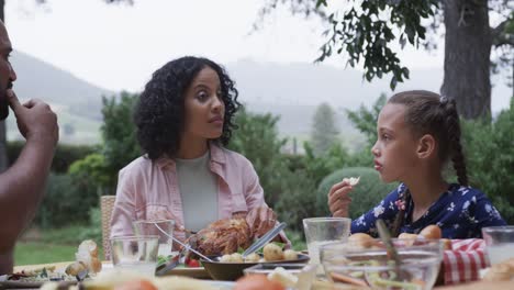 happy biracial parents and daughter eating meal at dinner table in garden, slow motion