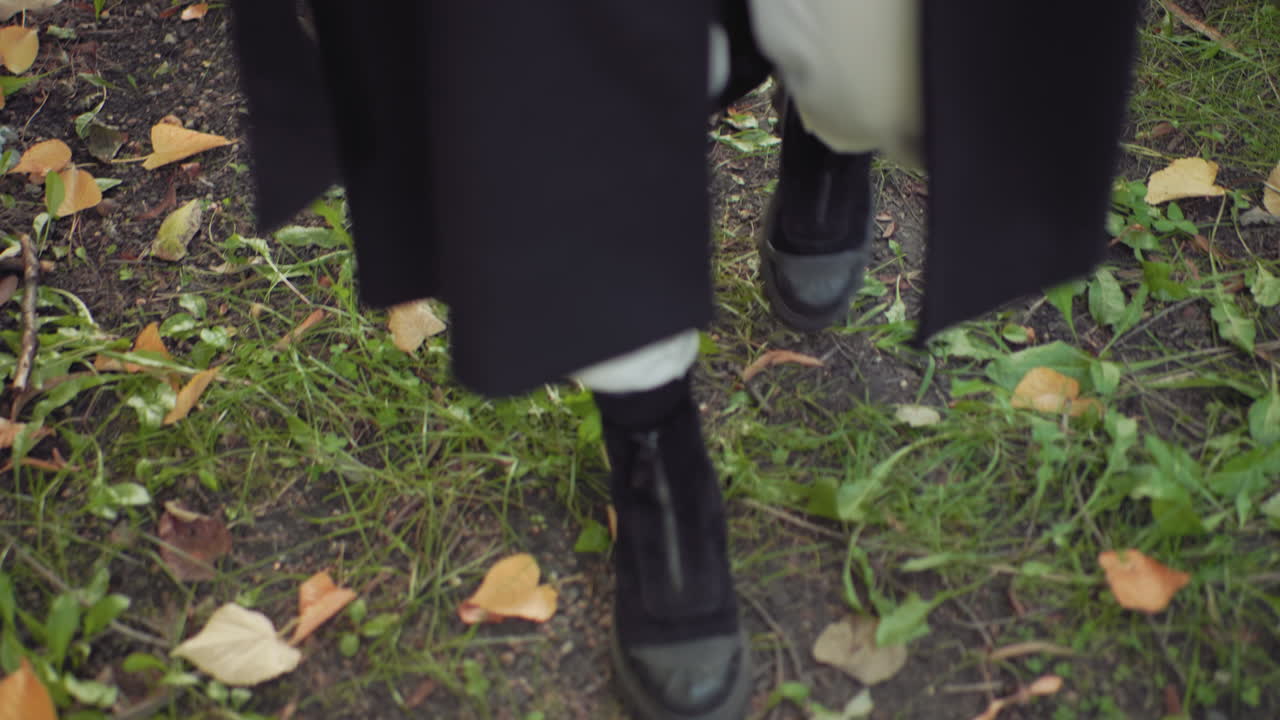 High angle view of lady in black boots walking on forest path, long coat swaying over white trousers, steps crunching dry leaves on earthy ground, calm pace during quiet autumn afternoon