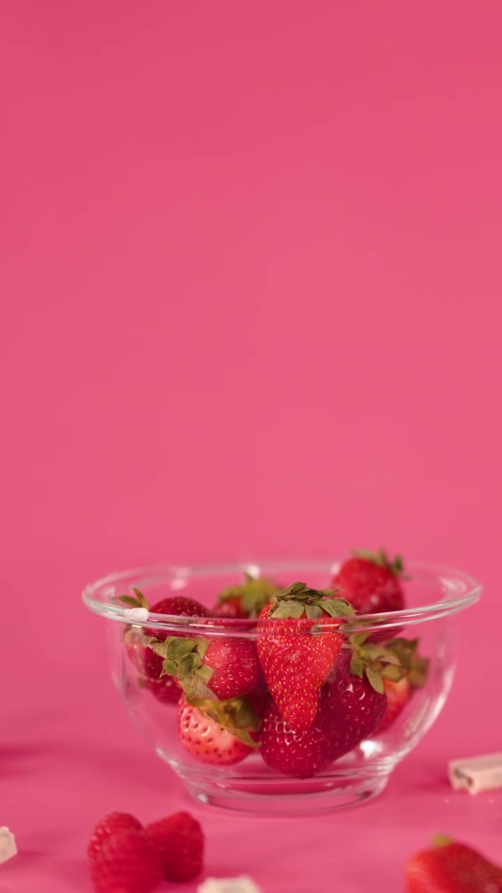 Strawberries, raspberries, and assorted sweets move into a glass bowl on a vibrant pink backdrop
