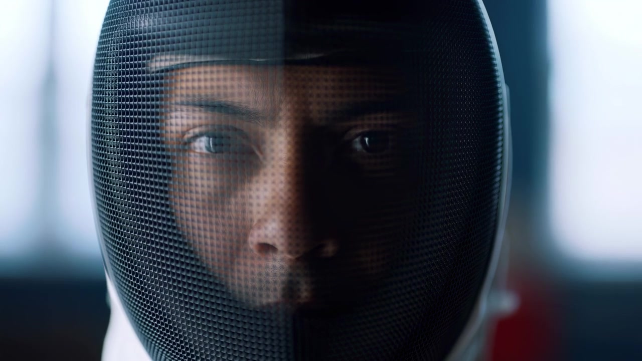 Tight head-on portrait of athlete wearing white fencing jacket and fine mesh mask, eyes visible through visor, lit by cool window light during indoor training