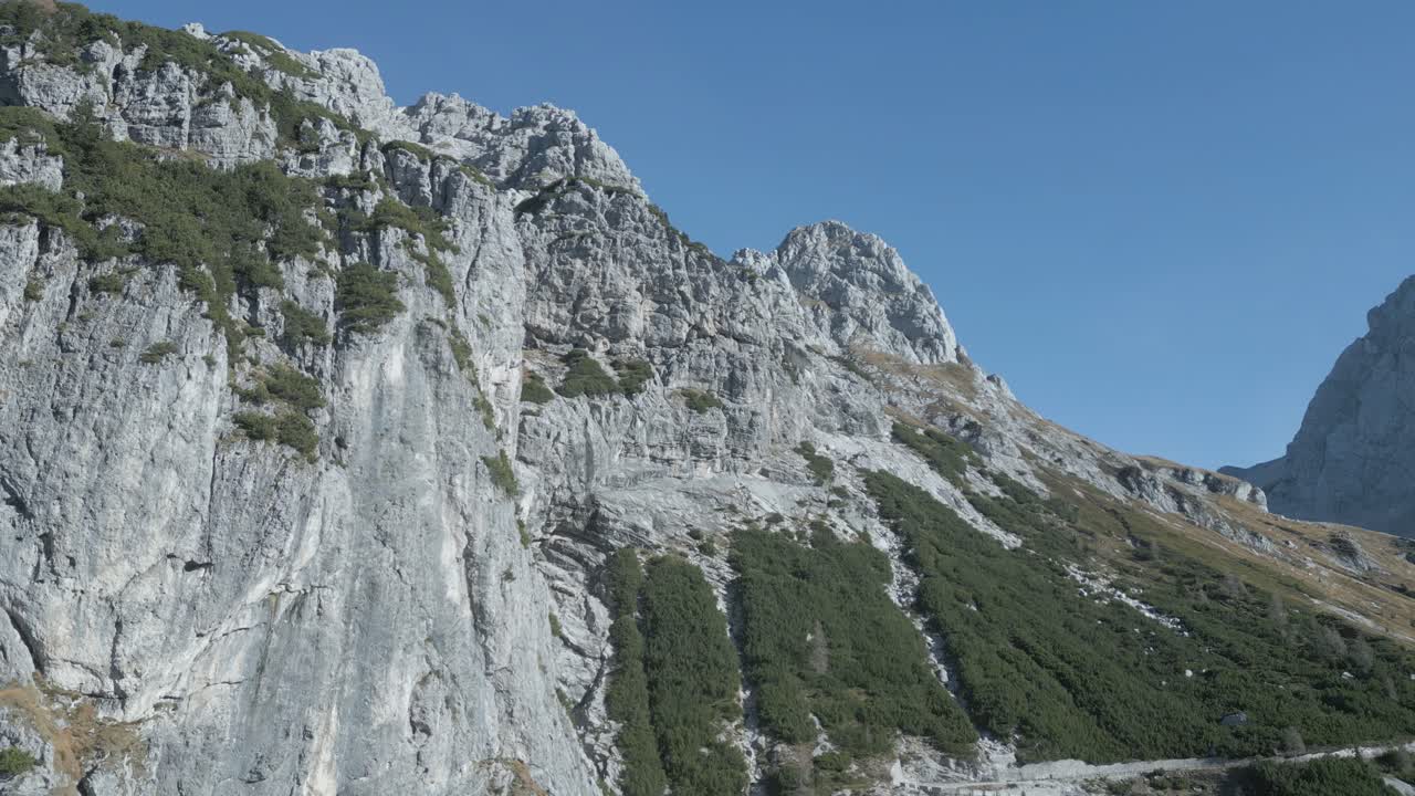 Drone view of Mangart Mountains, Slovenia: jagged peaks, rugged cliffs, lush green valleys, winding alpine roads, dramatic landscape, clear skies, Julian Alps, untouched natural beauty