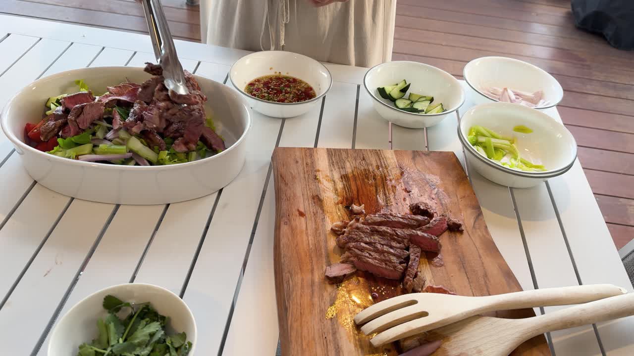 A person uses tongs to add sliced grilled beef to a large Thai salad bowl on a white table, surrounded by fresh ingredients and natural daylight