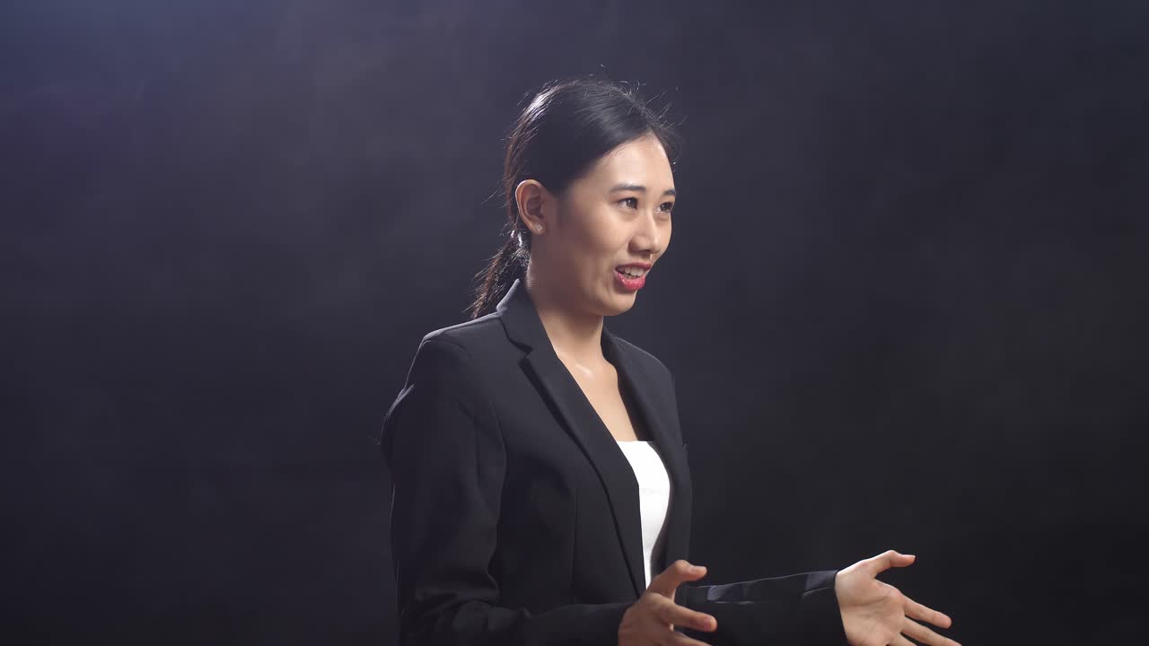 블랙 스크린 스튜디오에 서 있는 동안 엄지손가락을 올린 몸짓을 하는 비즈니스 슈트를 입은 미소 짓는 아시아 스피커 여성의 측면 시각