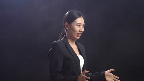 side view of smiling asian speaker woman in business suit showing thumbs up gesture while standing in the black screen studio