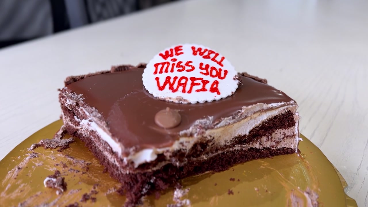 Primo piano di una torta d'addio a strati di cioccolato con un messaggio che dice "Ci mancherai Wafia" su una base dorata durante una festa in ufficio diurna. Messa a fuoco cinematografica selettiva sul dettaglio del dessert.