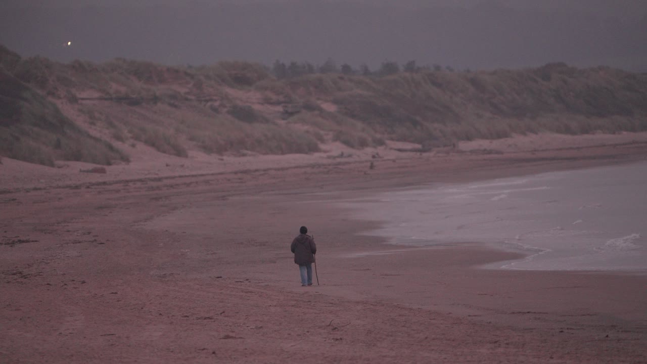 An elderly man walks alone along a quiet beach at sunset. Peaceful coastal evening scene evokes solitude, reflection, and the passing of time in nature
