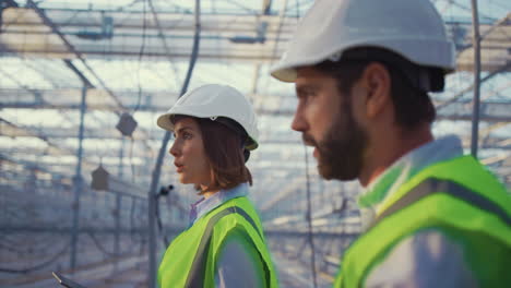 Portrait-uniform-engineers-talking-in-empty-glasshouse-analysing-manufacture.