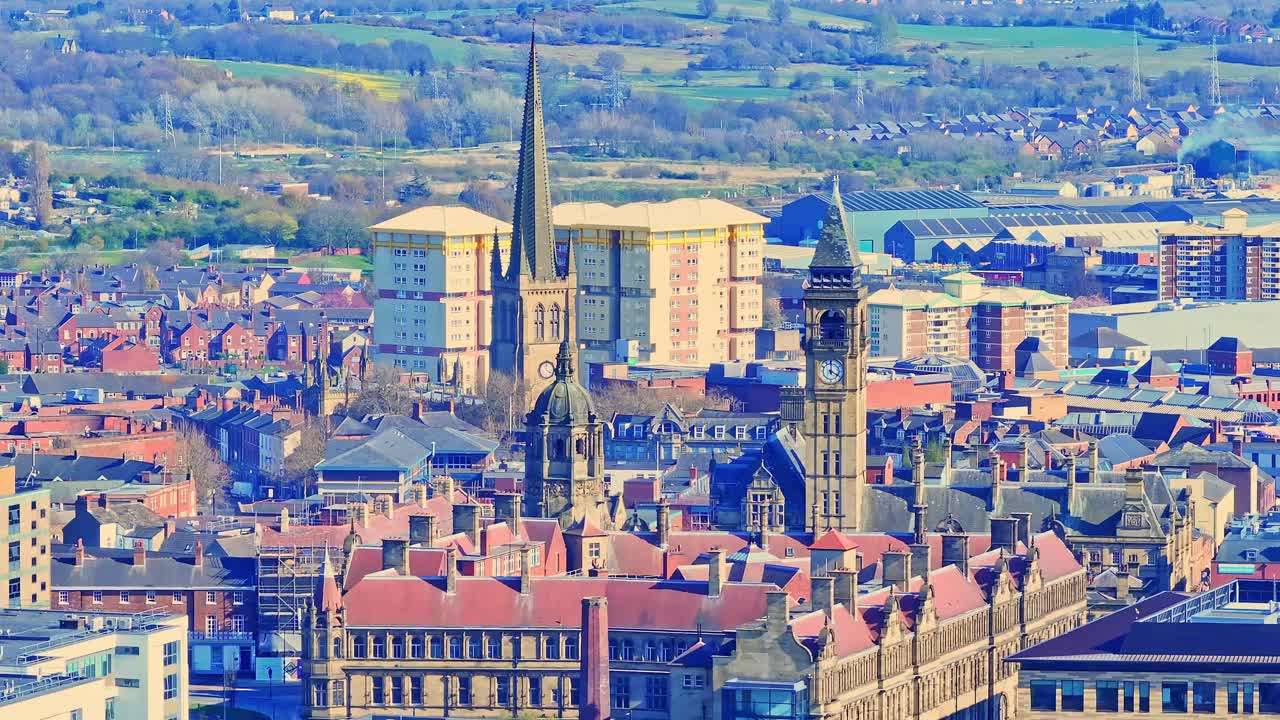 Aerial view of a cathedral church in Wakefield, England, surrounded by traditional brick housing, industrial buildings, with a clock tower by the dense urban cityscape. Under a sunny a day.