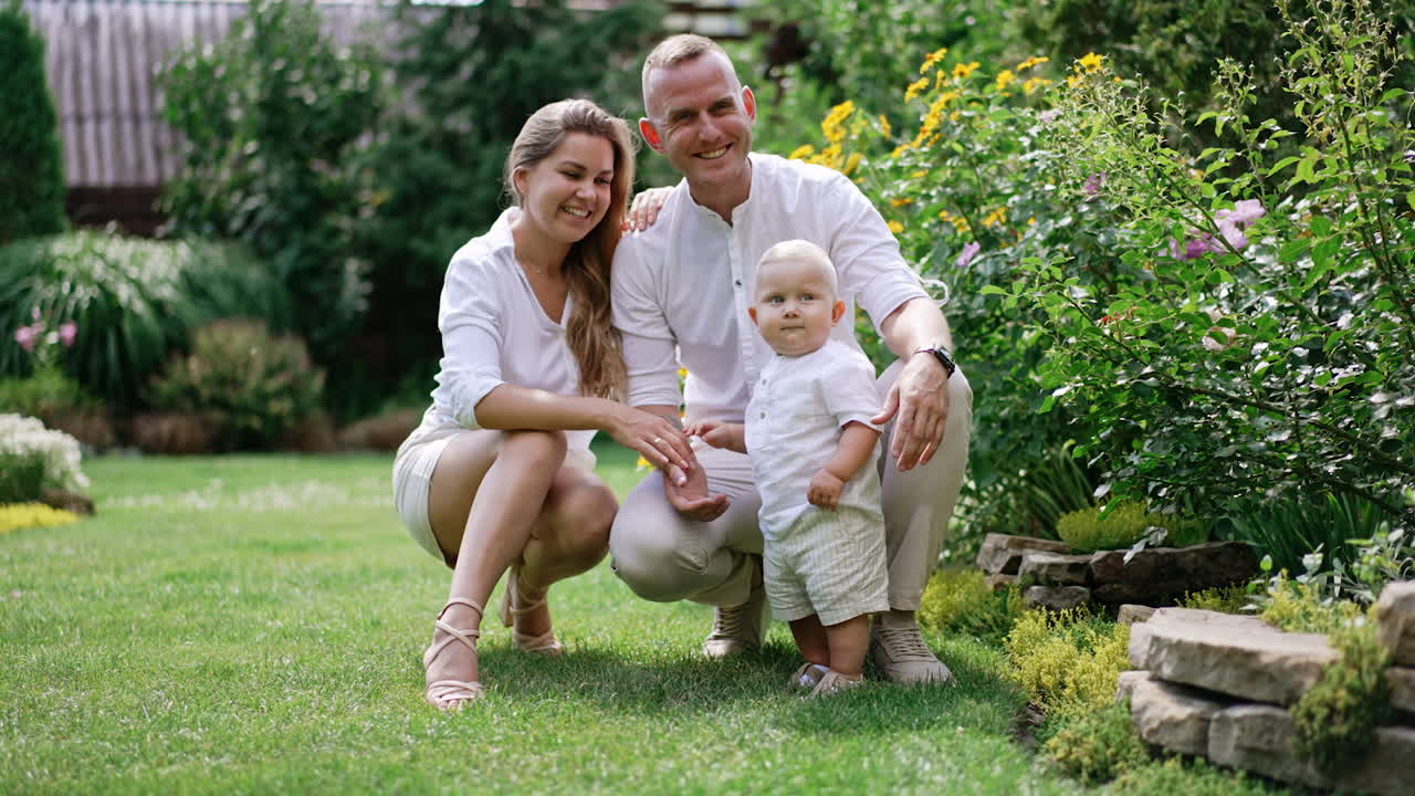Happy Caucasian parents smile to their little baby boy. Adorable child starts to dance. Beautiful garden at backdrop.