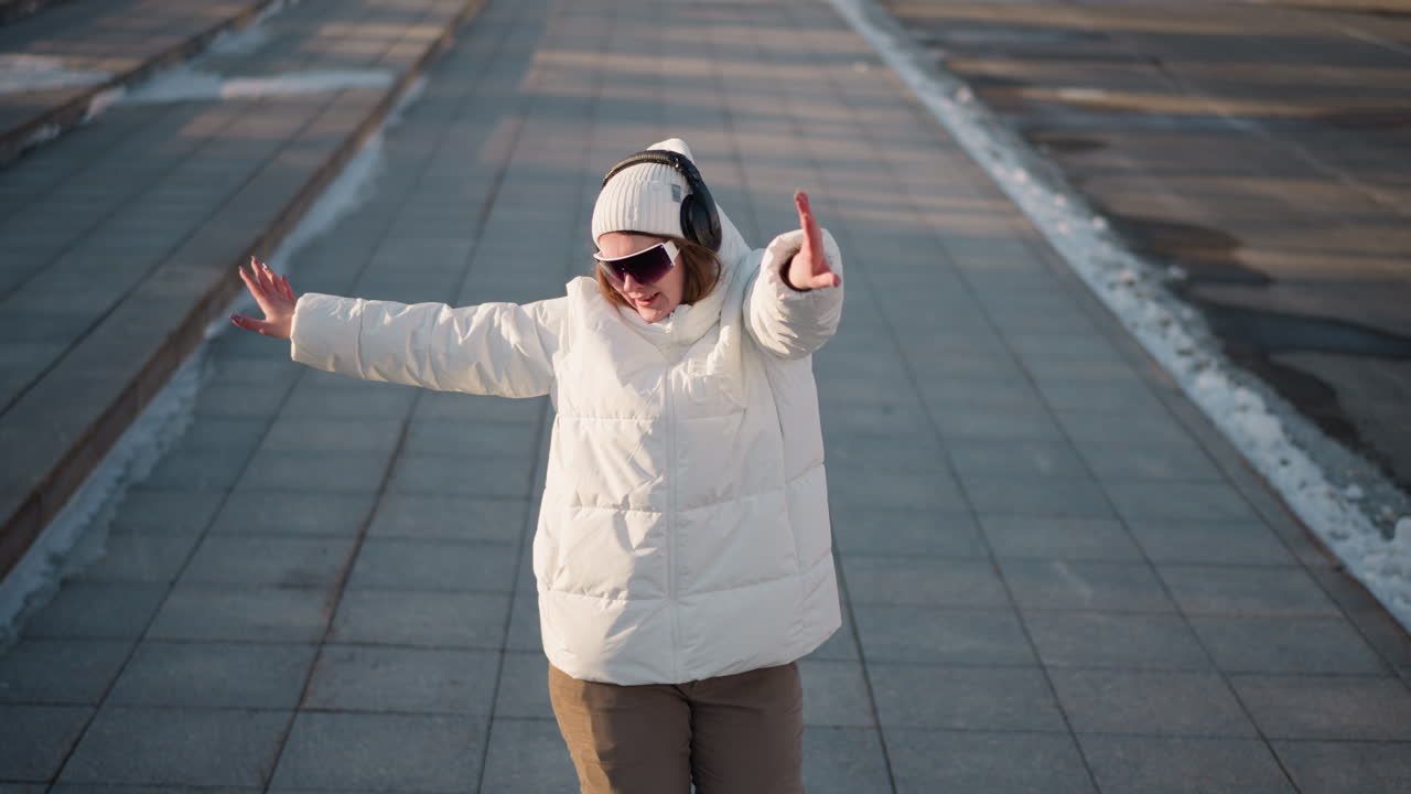 Joyful young lady dances and sings with headphones on, wearing white winter jacket and beanie, enjoying music while moving across sunlit snowy pavement in open city space during winter afternoon