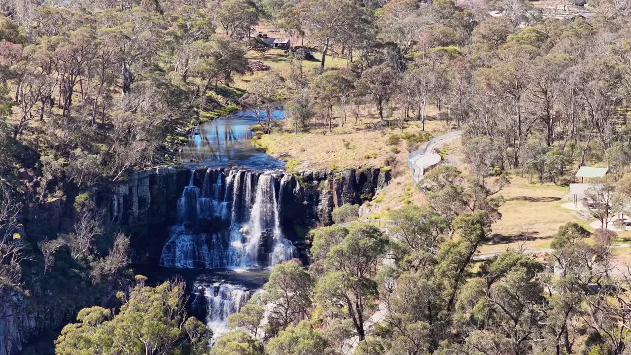 Aerial footage glides above Ebor Falls and a clifftop viewing platform in bright daylight, revealing forested surroundings and a winding path in rural New South Wales