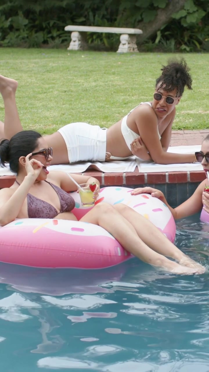 Vertical video: Sipping cocktail, two women in bikinis talking for fun by pink donut float poolside
