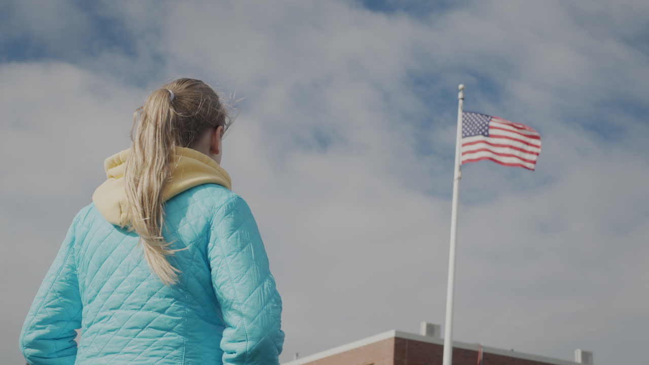 niño en edad escolar mirando la bandera de los estados unidos en el mástil, vista trasera