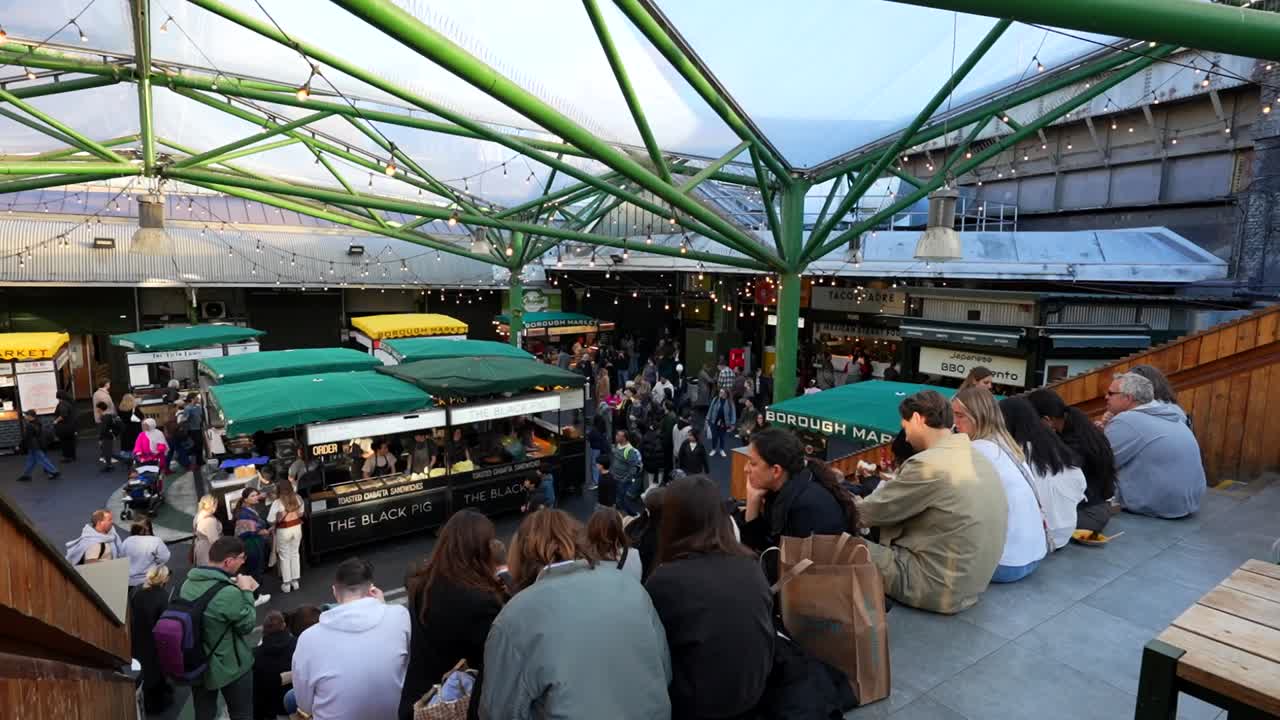 People enjoy meals on steps at Borough Market with lively food stalls and green steel structure above