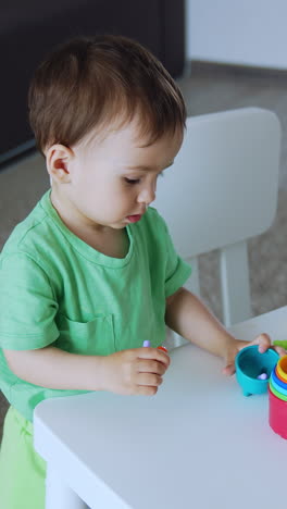 beautiful baby toy in green t-shirt stand near the desk. little kid is focused on the toys. top view. vertical video