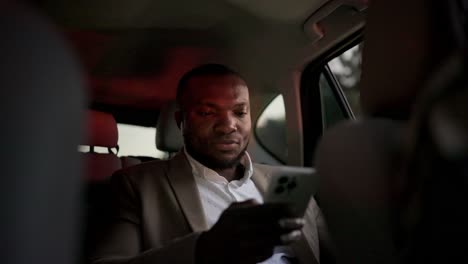 a confident man with black skin in a brown suit works on his white smartphone while driving on a business trip in the interior of a modern car