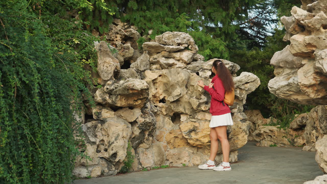 Young caucasian woman in red jacket exploring rocky garden wall amid trailing ivy, reaching toward stone crevices, stepping along gravel path, candid tourist and student persona, playful curiosity