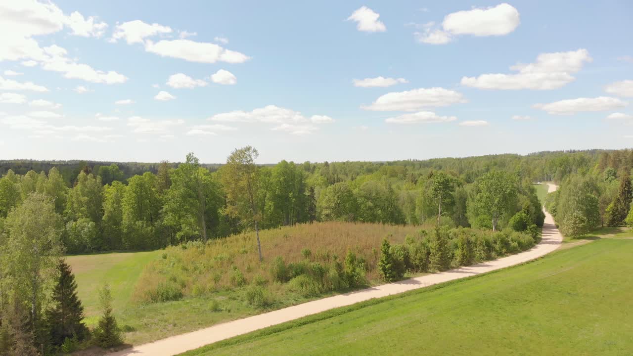 Drone flying up above northern European landscape with country road near vast forest, sunny summer day