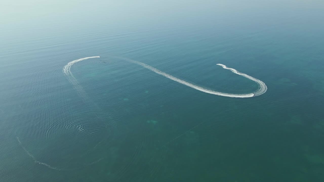 An aerial view captures a speedboat carving smooth, curved trails across a tranquil, deep blue sea. The wake forms a dynamic pattern on the otherwise still wate