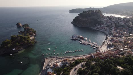 Barcos-Anclados-En-El-Muelle-De-La-Ciudad,-La-Gente-Camina-Por-El-Paseo-Marítimo,-Orbitando,-Vista-Aérea-De-La-Ciudad-Griega-Costera-De-Parga